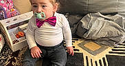 Alona is registered to the contest to win money with this photo: child, toddler, sitting, pacifier, bow_tie, cat_ears, white_shirt, gray_pants, barefoot, rug, living_room, couch, toys, toy_storage, backpack, clothing, pink_shoe, indoors, portrait, family_home