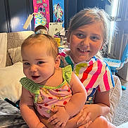 Alona is registered to the contest to win money with this photo: baby, child, sisters, toddler, smiling, portrait, indoors, seated, 抱抱, colorful_clothing, striped_dress, patterned_outfit, green_gingham, nursery, bedroom, toy, wall_art, furniture, family, happy