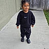 child, toddler, standing, outdoor, sidewalk, concrete, portrait, smiling, happy, athletic_wear, black_clothing, sneakers, sweatshirt, pants, curly_hair, hairstyle, backyard, fence, gate, residential