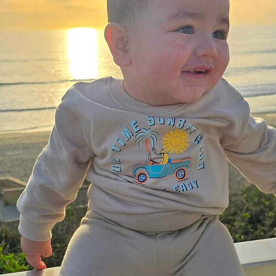 Koa joined the competition — help win amazing prizes! baby, beach, clothing, coast, face, grass, head, nature, outdoors, pants, person, photography, portrait, sea, shoreline, sky, sun, t_shirt, water, waterfront