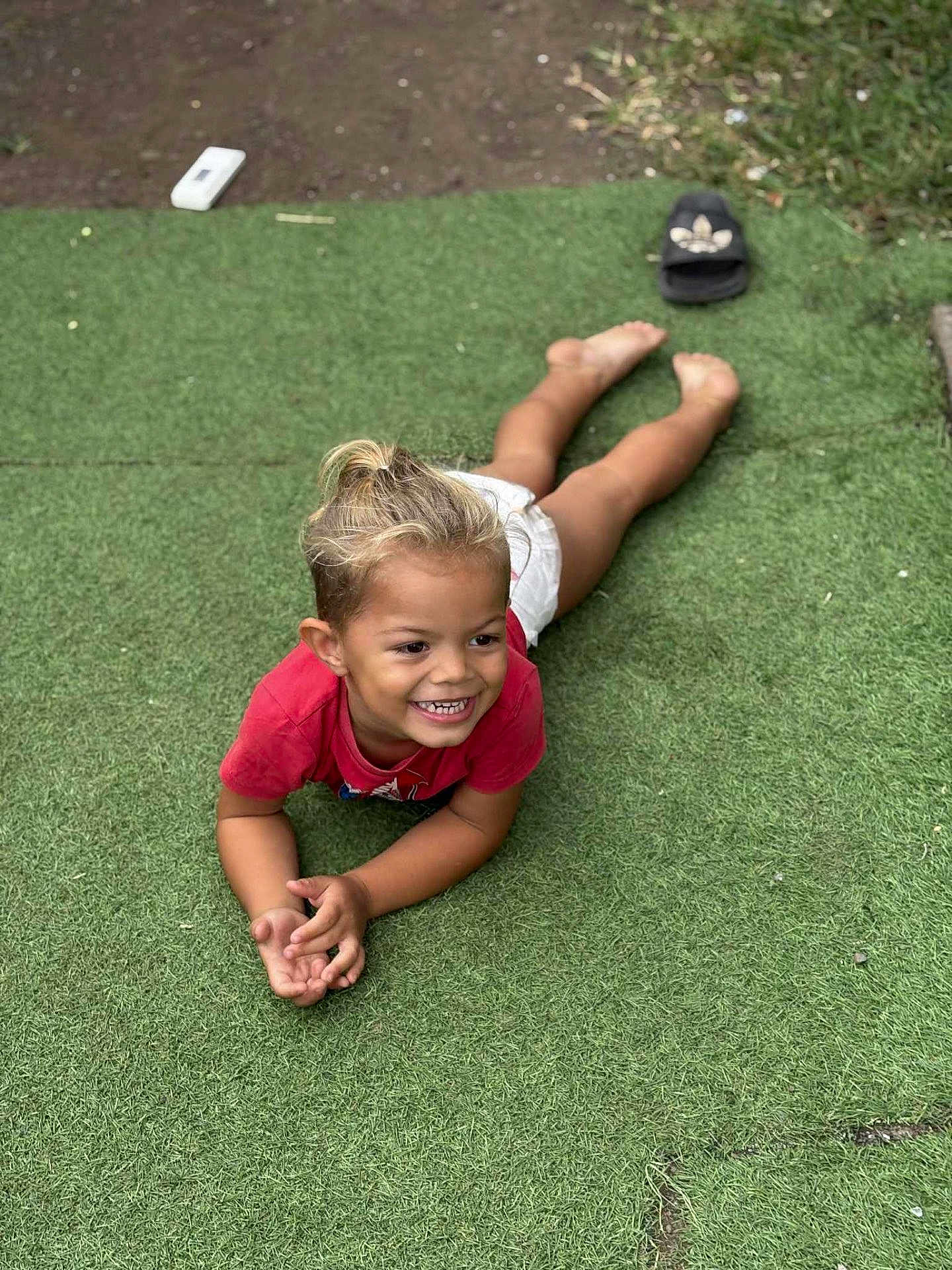 Kayden participe au concours pour gagner de l'argent avec cette photo : child, smiling, grass, artificial_turf, barefoot, red_shirt, shorts, outdoor, playful, happy, young, person, footwear, sandals, dirt, grass_patch, casual, summer, childhood, relaxed