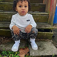 Bronx is registered to the contest to win money with this photo: toddler, child, curly_hair, white_shirt, camo_pants, white_sneakers, wooden_steps, outdoor, plants, fallen_leaves, concrete, greenery, curious_expression, casual_clothing, young_child, nature, daylight, sneakers, sitting, portrait