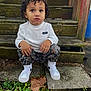 toddler, child, curly_hair, white_shirt, camo_pants, white_sneakers, wooden_steps, outdoor, plants, fallen_leaves, concrete, greenery, curious_expression, casual_clothing, young_child, nature, daylight, sneakers, sitting, portrait