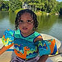 child, curly_hair, floatation_device, dinosaur_pattern, swimwear, boat, lake, water, trees, outdoor, summer, sunlight, nature, young_child, sitting, expression, water_safety, recreation, vacation, daytime