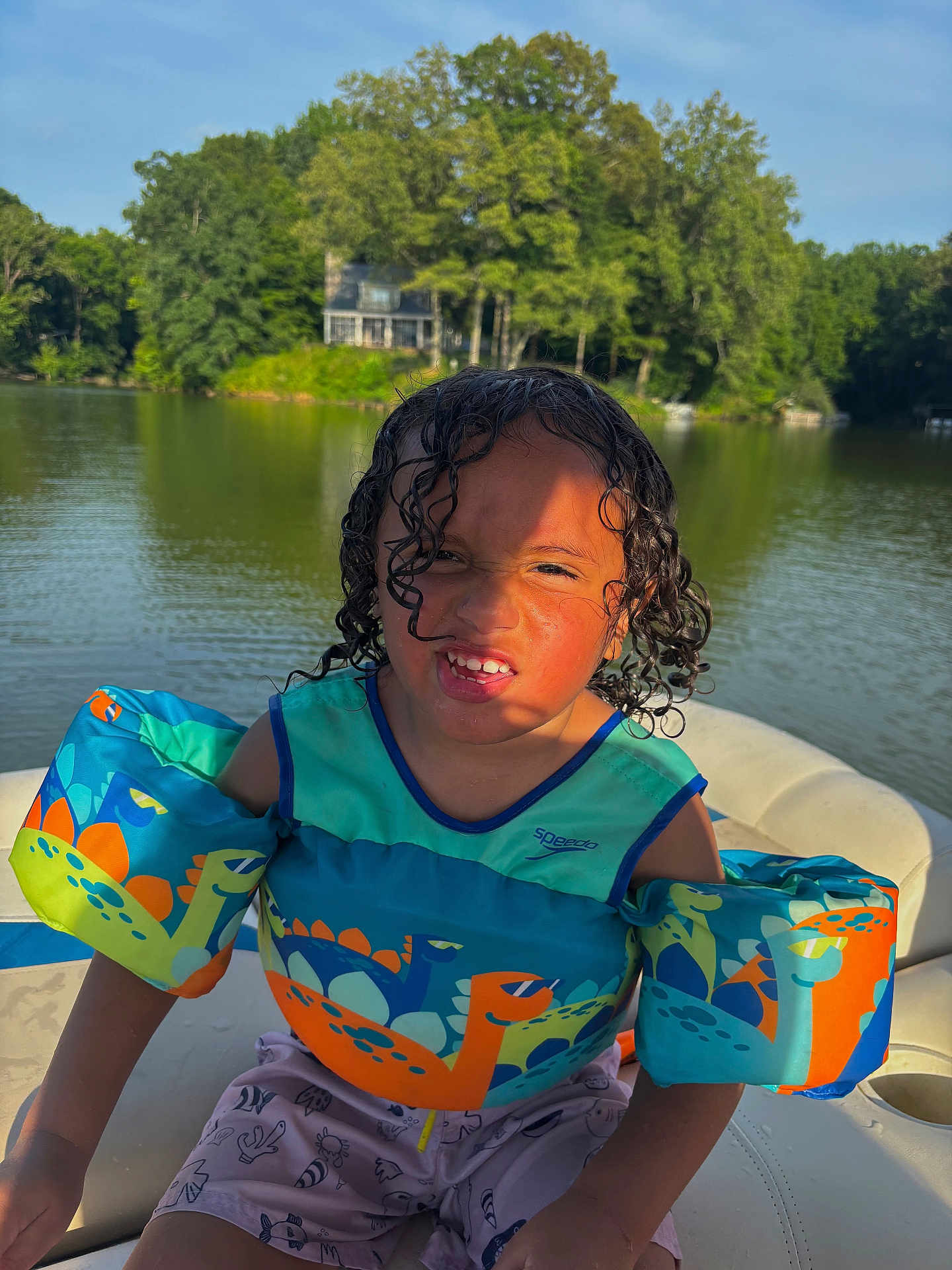 Bronx joined the competition — help win amazing prizes! child, boy, lake, water, boat, floatation_device, dinosaur_print, curly_hair, wet_hair, summer, outdoor, trees, house, sunlight, funny_face, sitting, life_jacket, nature, recreation, water_safety