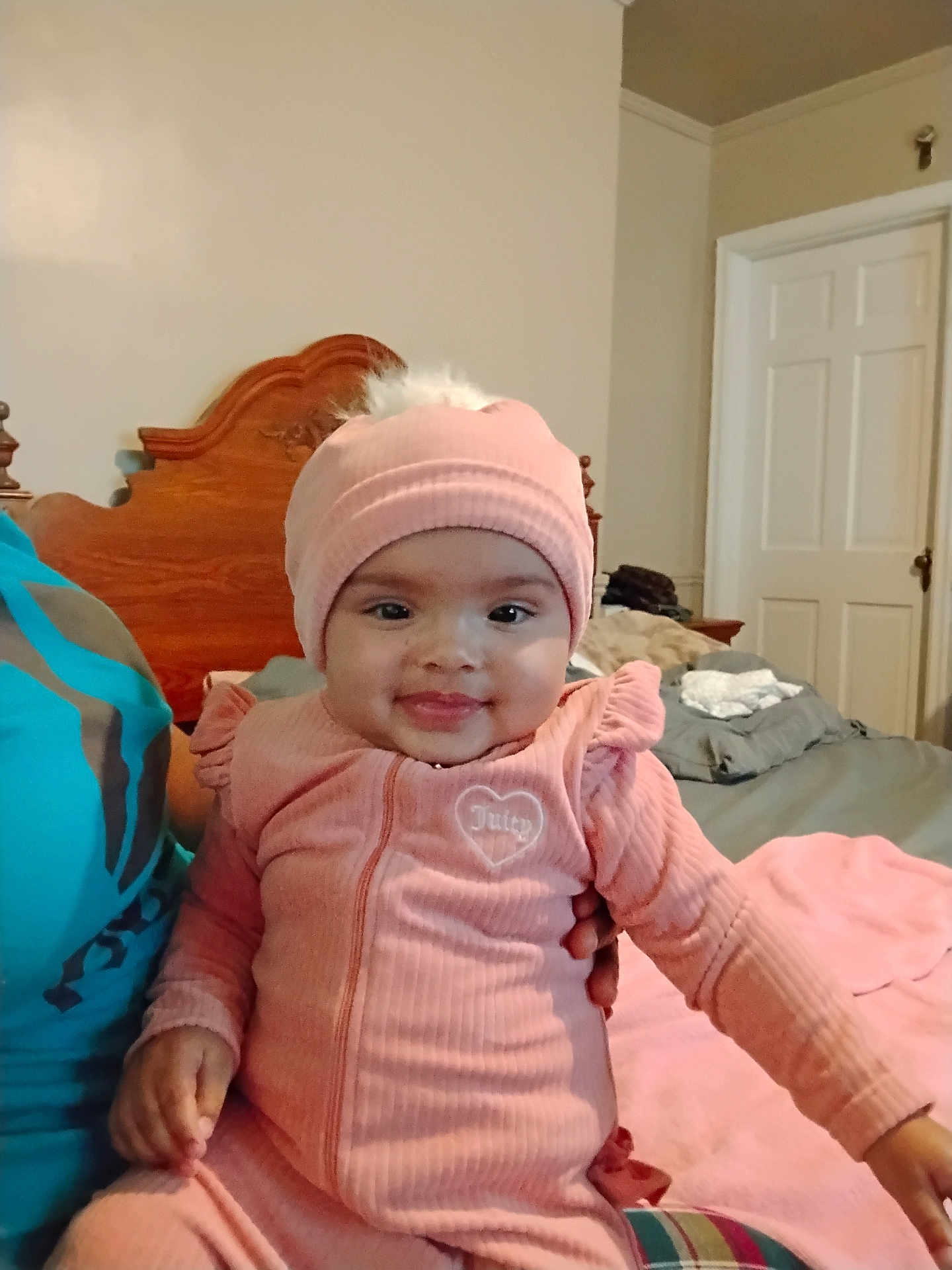 Akirra joined the competition — help win amazing prizes! baby, infant, pink_clothing, hat, smile, bedroom, bed, wooden_headboard, person, cute, indoor, child, joyful, warm_clothing, human, seated, cozy, happy, portrait, dressed