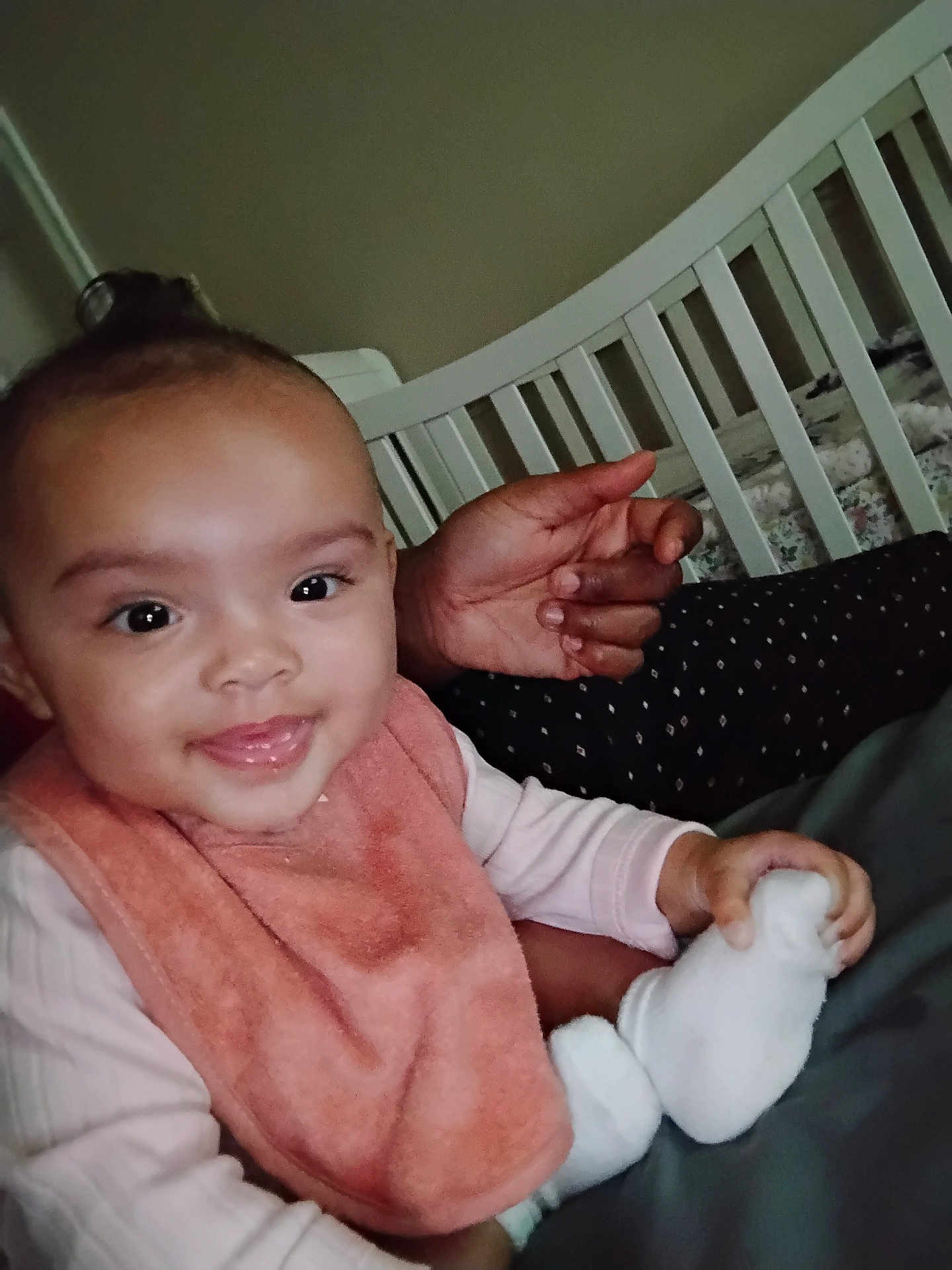 Akirra is registered to the contest to win money with this photo: baby, child, infant, smile, pink_bib, white_socks, adult_hand, crib, bedroom, furniture, person, happy, cute, indoors, portrait, holding, skin, face, clothing, baby_toys