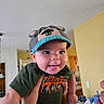 Callum is registered to the contest to win money with this photo: baby, child, cap, hat, indoors, person, smiling, tongue_out, cute, playful, hand, holding, face, clothing, kitchen, expression, infant, happy, young, portrait