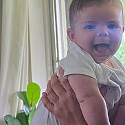 Callum is registered to the contest to win money with this photo: baby, infant, smiling, happy, person, child, hand, indoor, curtain, plant, greenery, face, skin, white_clothing, holding, portrait, toddler, expression, cute, young
