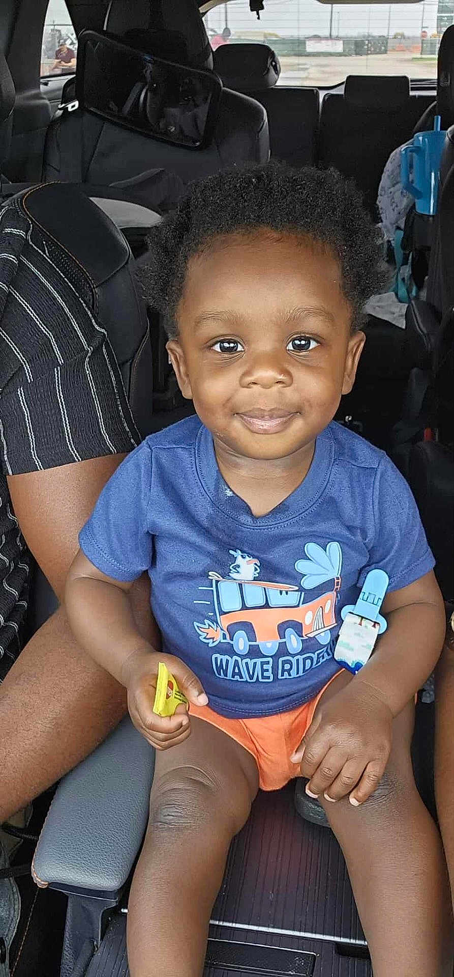 Haysen is registered to the contest to win money with this photo: toddler, child, smiling, blue_tshirt, orange_shorts, car_seat, inside_car, pacifier, snack, hands, legs, curly_hair, person, seat, window, daylight, vehicle, happy, casual_clothing, portrait