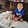 Chase is registered to the contest to win money with this photo: boy, dog, couch, blanket, bowl, cereal, spoon, pillow, flower_pattern, home_decor, child, pet, indoor, smile, casual_clothing, brown_furniture, white_blanket, relaxed, cute, happy