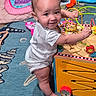 baby, child, toy, bead_maze, play, colorful, rug, indoors, smile, standing, blanket, furniture, wooden_toy, floor, person, cute, happy, playroom, foot, hand