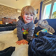 Addam participe au concours pour gagner de l'argent avec cette photo : blanket, blond_hair, bottle, carpet, chandelier, child, couch, furniture, hands, hoodie, living_room, playful, plush, portrait, smiling, stone_wall, sunlight, toddler, toy, window