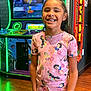 child, girl, arcade, arcade_game, smiling, pink_clothing, princess_print, tooth_gap, indoor, wooden_floor, gaming, bright_colors, neon_lights, happy, short_sleeve, standing, casual_wear, entertainment, fun, portrait