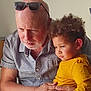 elderly_man, child, indoor, curly_hair, yellow_shirt, sunglasses, gentle_touch, family, intense_gaze, human, portrait, candid, affection, casual_clothing, beard, short_sleeves, light_skin, brown_skin, tender_moment