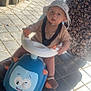 baby, child, hat, toy, ride_on_toy, outdoor, sunlight, pavement, person, floral_dress, hand, scooter, straps, footwear, curious, expression, playful, summer, happy, walking_assist