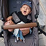 baby, stroller, cap, yawning, toy, urban, pavement, stone_steps, outdoor, child, black_clothing, gray_stroller, foot, hand, seatbelt, wheel, fabric, cute, infant, person