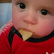 Hudson is registered to the contest to win money with this photo: baby, infant, child, portrait, close_up, face, eyes, smiling, curious, snack, food, eating, red_shirt, clothing, indoor, home, seated, adorable, photography, black_background