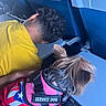 Cookie is registered to the contest to win money with this photo: service_dog, dog, pet, animal, person, yellow_shirt, airplane_seat, travel, interior, seat_back, tray_table, curly_hair, hand, blue_background, canine, fur, harness, resting, companion, indoor