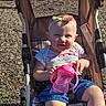 baby, child, stroller, sippy_cup, headband, bow, outdoor, playground, sunlight, wood_chips, clothing, shorts, smile, person, seat, mesh, fabric, hand, legs, happy