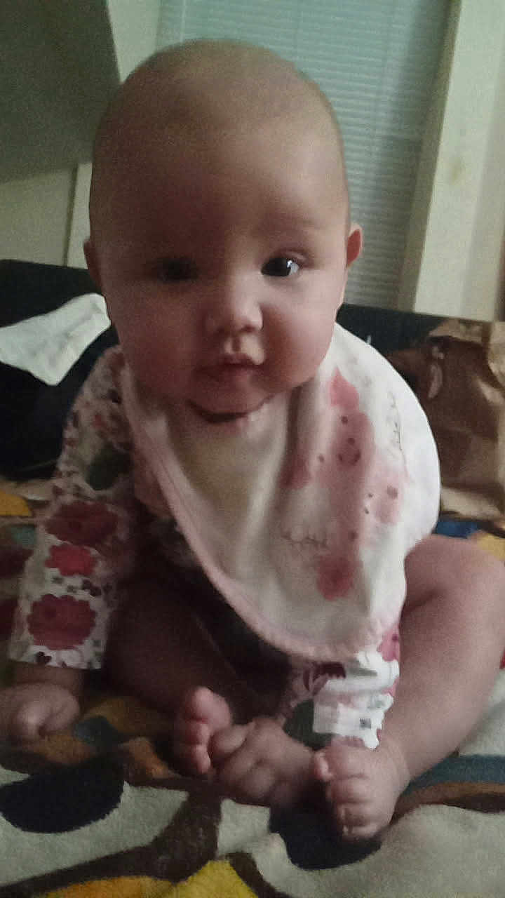 Roselyne joined the competition — help win amazing prizes! baby, infant, child, bib, floral_clothing, blanket, feet, face, sitting, indoors, curious, cute, portrait, person, head, eyes, hands, soft_lighting, young, front_view