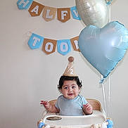 Noah is registered to the contest to win money with this photo: baby, balloons, birthday, birthday_banner, blue_clothing, celebration, child, curly_hair, decorations, happy, high_chair, indoor, infant, one_year_old, party_hat, photography, portrait, sitting, smiling, wall