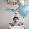 baby, balloons, birthday, birthday_banner, blue_clothing, celebration, child, curly_hair, decorations, happy, high_chair, indoor, infant, one_year_old, party_hat, photography, portrait, sitting, smiling, wall