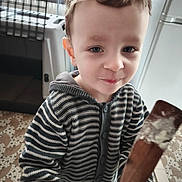 Aurélien a rejoint le concours — aidez-le/la à gagner de superbes lots ! child, toddler, face, smile, striped_sweater, hoodie, wooden_spatula, batter, kitchen, refrigerator, pet_crate, tiled_floor, home_interior, candid, portrait, hair, eyes, hand, countertop, casual