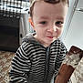 child, toddler, face, smile, striped_sweater, hoodie, wooden_spatula, batter, kitchen, refrigerator, pet_crate, tiled_floor, home_interior, candid, portrait, hair, eyes, hand, countertop, casual