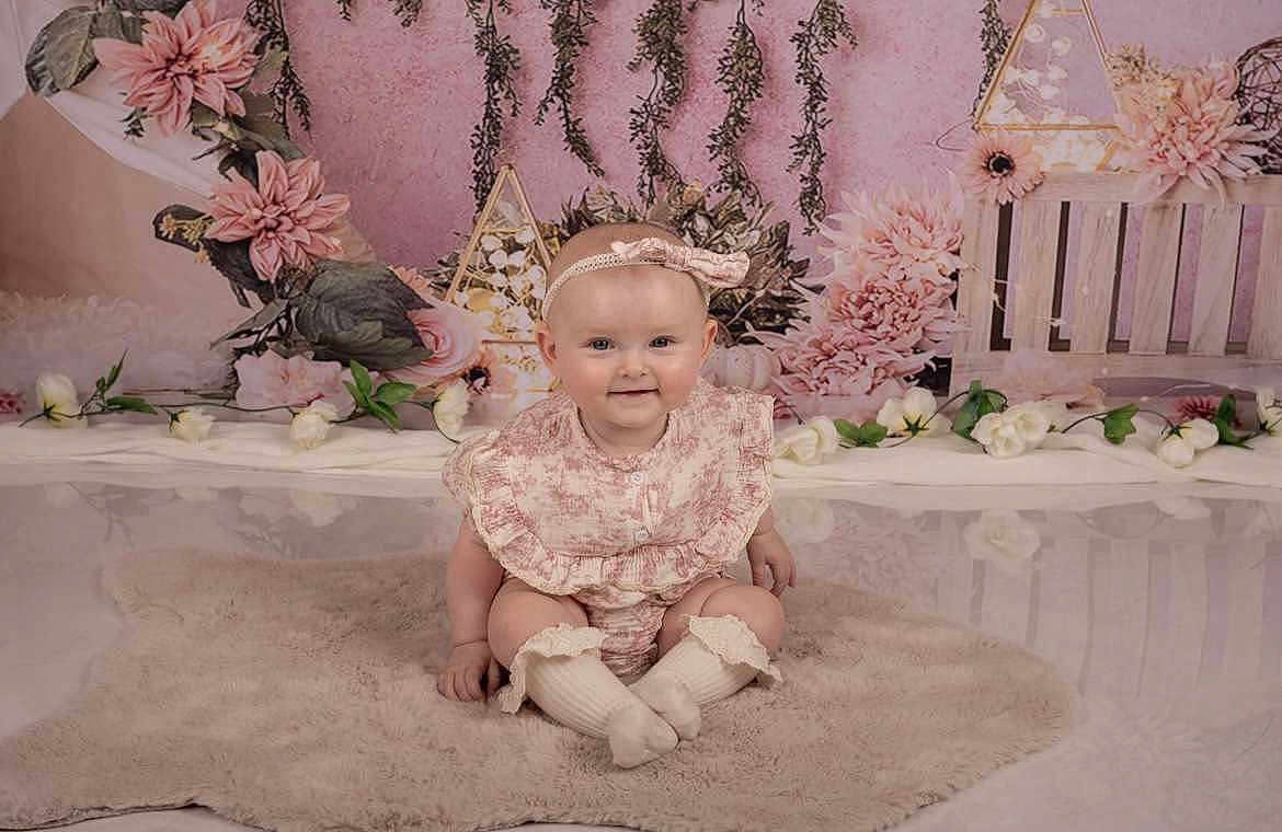 Romane participe au concours pour gagner de l'argent avec cette photo : baby, child, infant, smiling, sitting, headband, floral_outfit, pink, flowers, greenery, rug, indoor, portrait, cute, happy, decor, soft_texture, female, young_child, studio