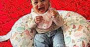 Emerie joined the competition — help win amazing prizes! baby, bed, child, clothing, cute, dinosaur_pattern, happy, headband, indoor, infant, jeans, joyful, laughing, person, pillow, portrait, red_bedspread, seated, smiling, sneakers