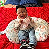 baby, bed, child, clothing, cute, dinosaur_pattern, happy, headband, indoor, infant, jeans, joyful, laughing, person, pillow, portrait, red_bedspread, seated, smiling, sneakers