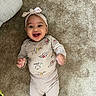Emerie is registered to the contest to win money with this photo: apparel, baby, baby_girl, carpet, child, clothing, cute, face, happy, head, headband, indoor, infant, lying_down, onesie, person, playful, portrait, smiling, young_child