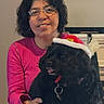Perrita Gabby joined the competition — help win amazing prizes! woman, dog, glasses, pink_sweater, black_dog, santa_hat, indoor, fireplace, television, smile, seated, holding, pet, festive, casual_clothing, home, technology, phone_screen, portrait, happy
