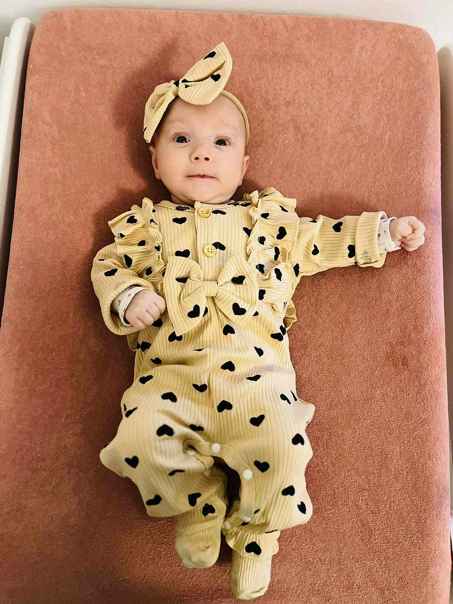 Ava participe au concours pour gagner de l'argent avec cette photo : baby, infant, onesie, headband, bow, heart_pattern, changing_pad, mattress, pink_background, cute, portrait, lying_down, hands, feet, ruffles, buttons, newborn, eyes, clothing, smile
