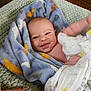 baby, blanket, smiling, infant, face, hand, nail_polish, blue_blanket, patterned_blanket, soft_texture, green_surface, wrapped, cute, child, skin, happy, indoors, closeup, person, toddler