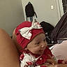 baby, infant, smile, red_hat, bow, floral_outfit, hand, indoor, person, child, furniture, backpack, wall, door, bed, happy, clothing, headwear, skin, portrait