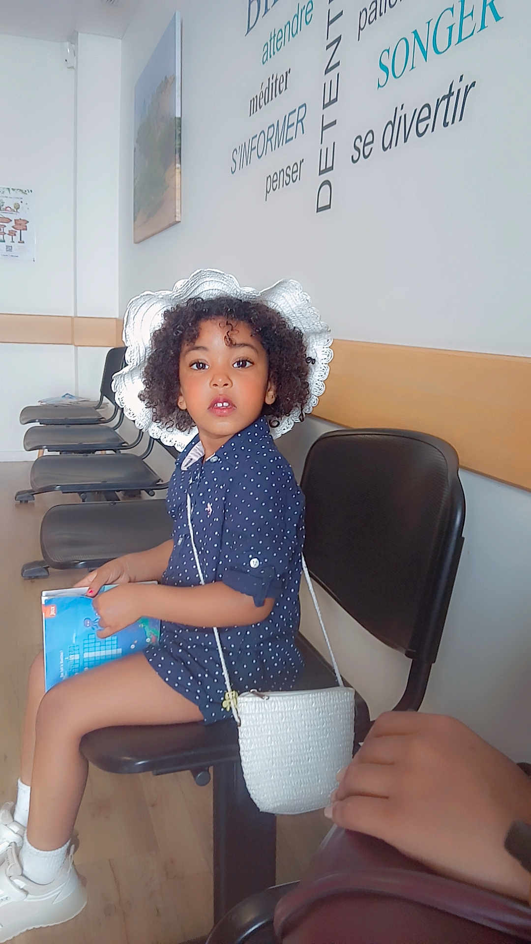 Myra a rejoint le concours — aidez-le/la à gagner de superbes lots ! child, girl, curly_hair, white_hat, waiting_room, book, purse, chair, polka_dot_dress, socks, white_shoes, wooden_floor, wall_art, empty_chairs, indoor, portrait, sitting, curious, person, casual
