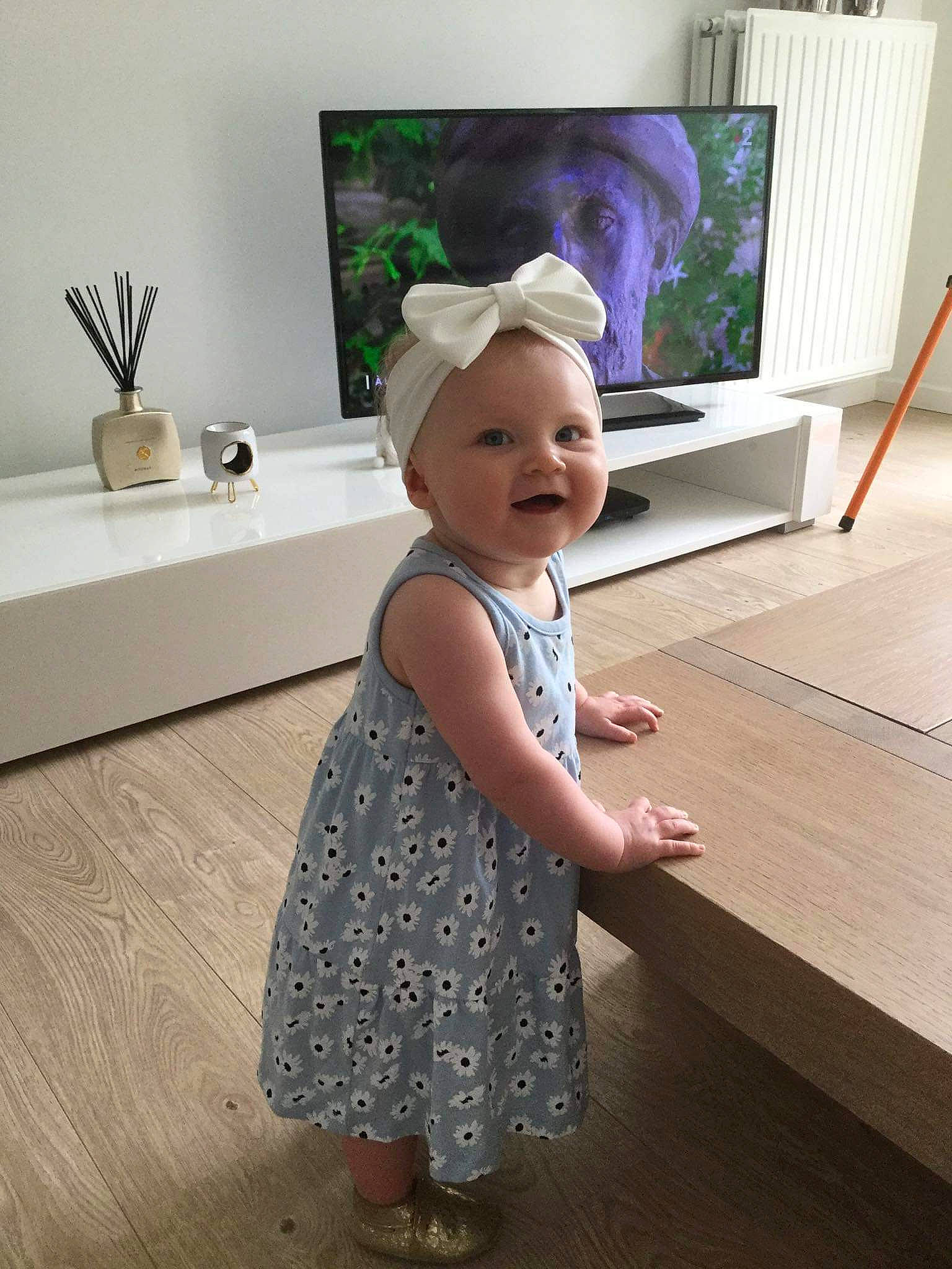 Lya a rejoint le concours — aidez-le/la à gagner de superbes lots ! baby, baby_toddler_clothing, child, day_dress, flooring, fun, happy, hardwood, hat, living_room, one_piece_garment, pattern, person, pink, sleeve, smile, standing, television, television_set, toddler