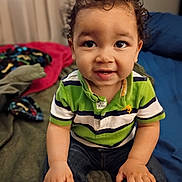 Ezra joined the competition — help win amazing prizes! toddler, child, curly_hair, green_shirt, striped_shirt, jeans, indoors, bed, blanket, smiling, portrait, cute, baby, person, hands, sitting, curious, young_child, cozy, casual_clothing