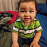 toddler, child, curly_hair, green_shirt, striped_shirt, jeans, indoors, bed, blanket, smiling, portrait, cute, baby, person, hands, sitting, curious, young_child, cozy, casual_clothing