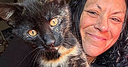 Sam is registered to the contest to win money with this photo: cat, tortoiseshell_cat, woman, person, selfie, portrait, smiling, indoor, pet, animal, feline, whiskers, green_eyes, fur, black_fur, orange_fur, nose_ring, black_clothing, close_up, home