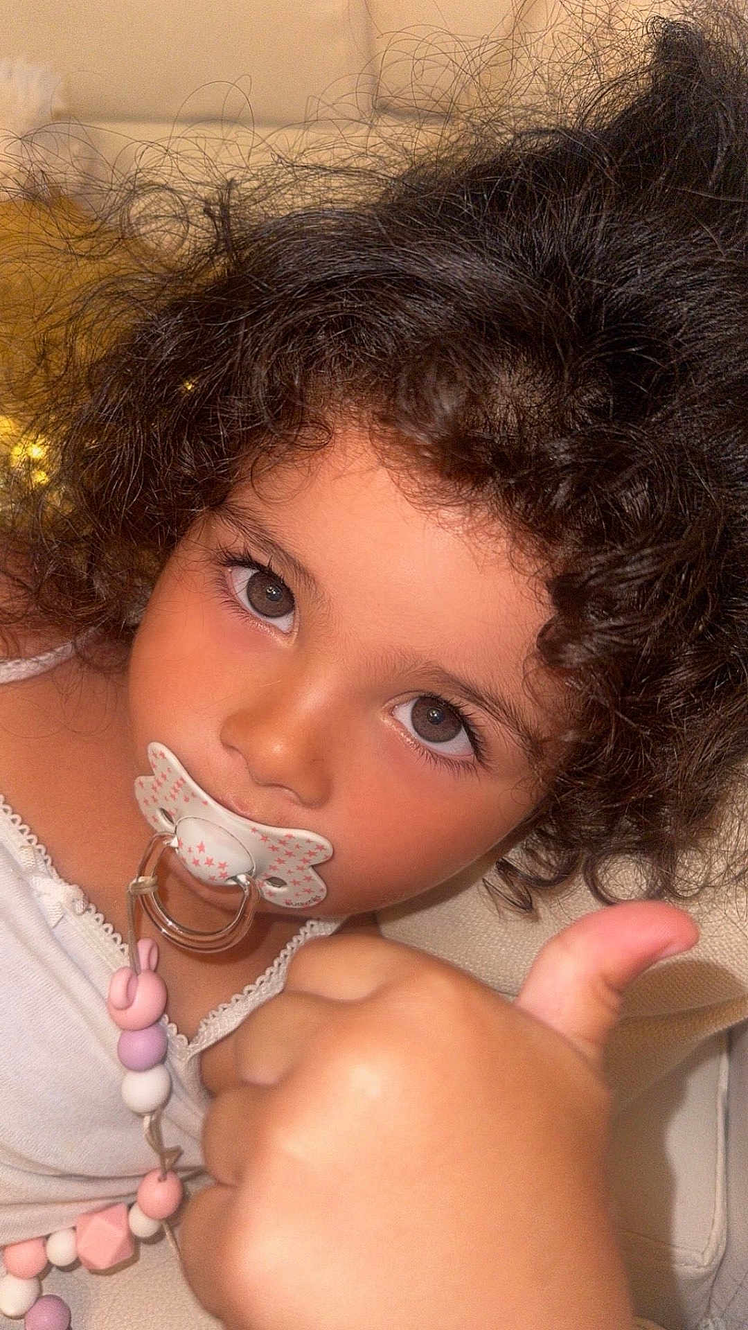 Numidia a rejoint le concours — aidez-le/la à gagner de superbes lots ! child, toddler, pacifier, thumbs_up, curly_hair, close_up, face, expression, indoor, skin, cute, young_child, person, portrait, baby_accessory, hand, seated, clothing, beads, soft_lighting