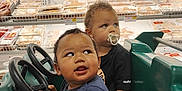 Justin is registered to the contest to win money with this photo: toddler, child, toy_car, grocery_store, packaged_meat, shopping_cart, pacifier, curly_hair, blue_shirt, black_shirt, steering_wheel, supermarket, indoor, baby, shopping, food, aisle, cute, play, vehicle