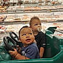 toddler, child, toy_car, grocery_store, packaged_meat, shopping_cart, pacifier, curly_hair, blue_shirt, black_shirt, steering_wheel, supermarket, indoor, baby, shopping, food, aisle, cute, play, vehicle