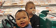Maverick is registered to the contest to win money with this photo: black_shirt, blue_shirt, car, child, curly_hair, cute, grocery_store, happy, indoor, meat_section, pacifier, packaged_food, playful, shopping_cart, smiling, steering_wheel, supermarket, toddler, toy_car, two_children