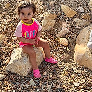 Julieta is registered to the contest to win money with this photo: casual_clothing, child, curly_hair, daylight, ground, happy_text, nature, outdoor, pebbles, person, pink_shirt, pink_shoes, rock, rocks, shoes, sitting, small_rocks, sunlight, toddler, young_child