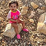 casual_clothing, child, curly_hair, daylight, ground, happy_text, nature, outdoor, pebbles, person, pink_shirt, pink_shoes, rock, rocks, shoes, sitting, small_rocks, sunlight, toddler, young_child