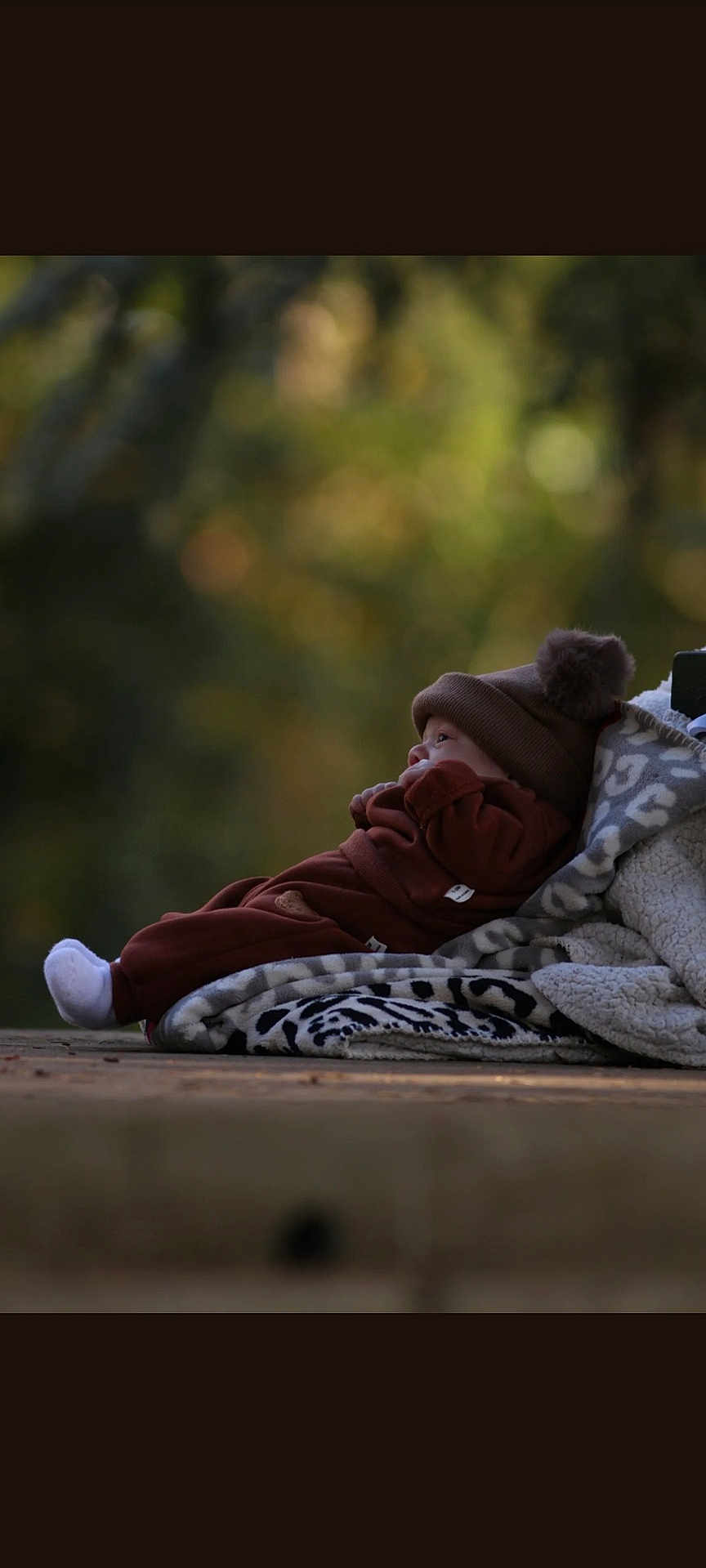 Noë participe au concours pour gagner de l'argent avec cette photo : baby, infant, hat, brown_clothing, blanket, pom_pom, outdoor, nature, wooden_surface, cozy, warm_clothes, soft_texture, side_view, portrait, child, cute, relaxed, curious, small, resting