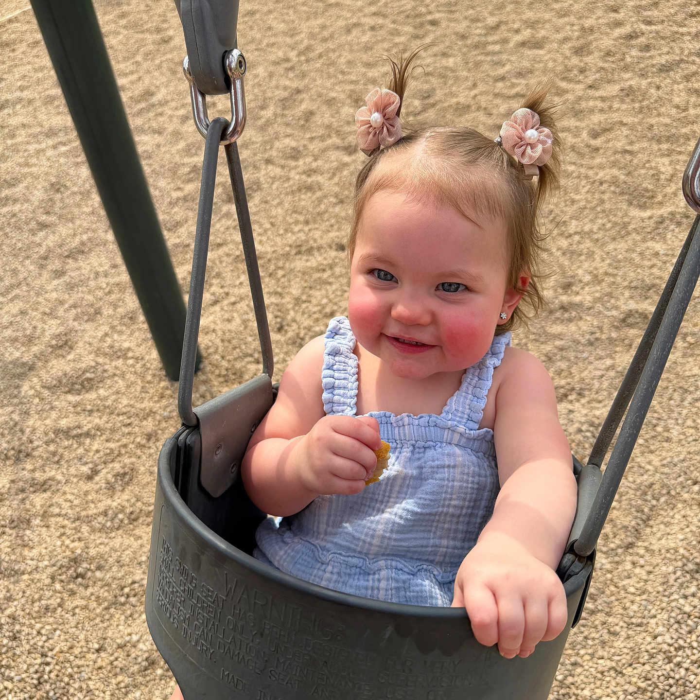 Aubrey is registered to the contest to win money with this photo: baby, child, toddler, swing, playground, outdoor, park, smiling, happy, portrait, face, pigtails, hair_bows, blue_outfit, romper, snack, sitting, play_equipment, sunlight, pebbles